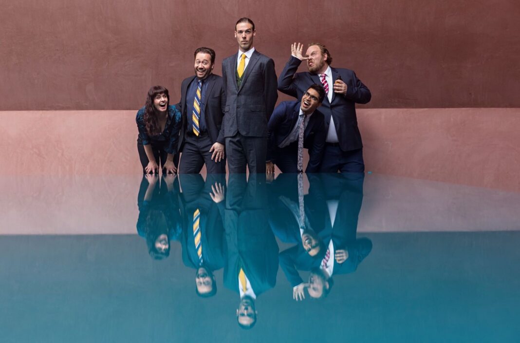 Five actors in suits striking poses near reflection pool