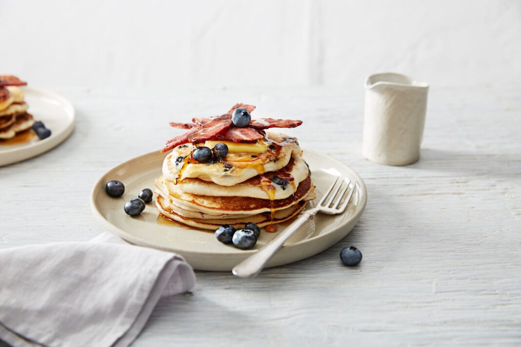 Stack of pancakes topped with blueberries, maple syrup and bacon strips on a white plate.