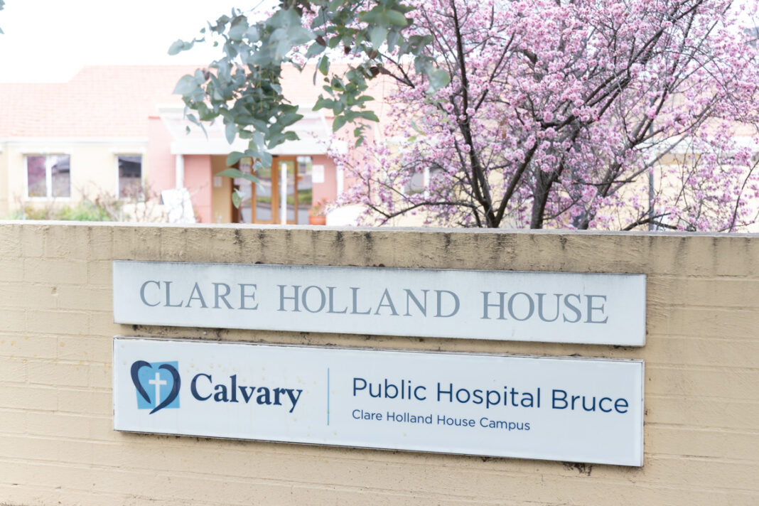 Sign that reads 'Clare Holland House' in front of hospice building and trees in blossom