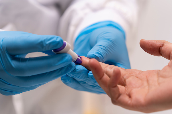 two hands wearing blue disposable hygiene gloves conducting finger prick blood test on another person's right hand