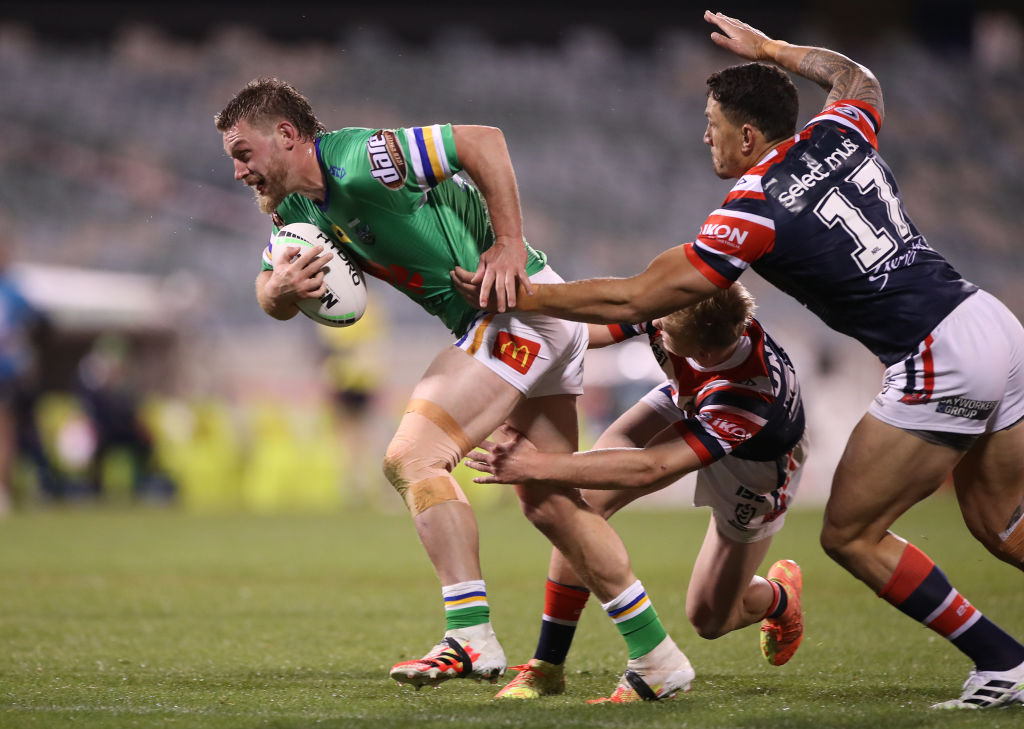 Rugby league player in green jersey making his way downfield with two players in black and red jerseys trying to tackle him