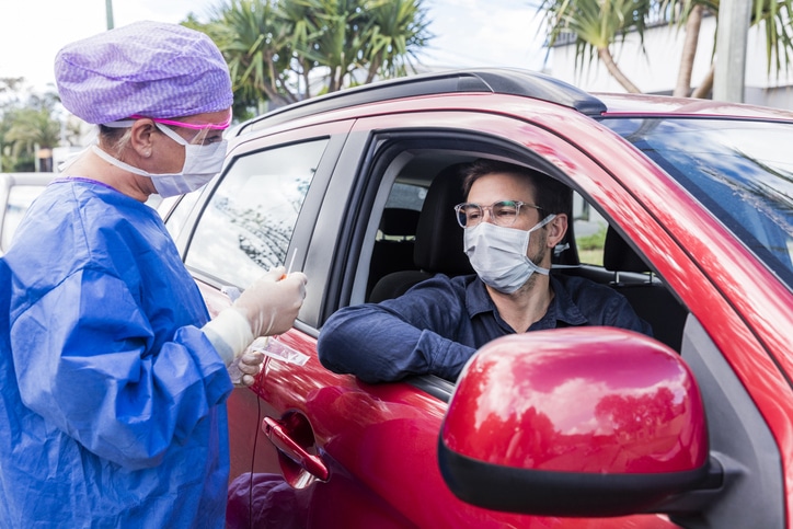 A female doctor in a protective suit taking a nasal swab from a man in a red car for possible coronavirus infection