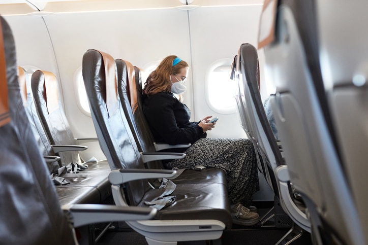 Side view of solitary Caucasian 17 year old girl wearing casual clothing and protective mask checking smart phone while flying on airplane in time of COVID-19.