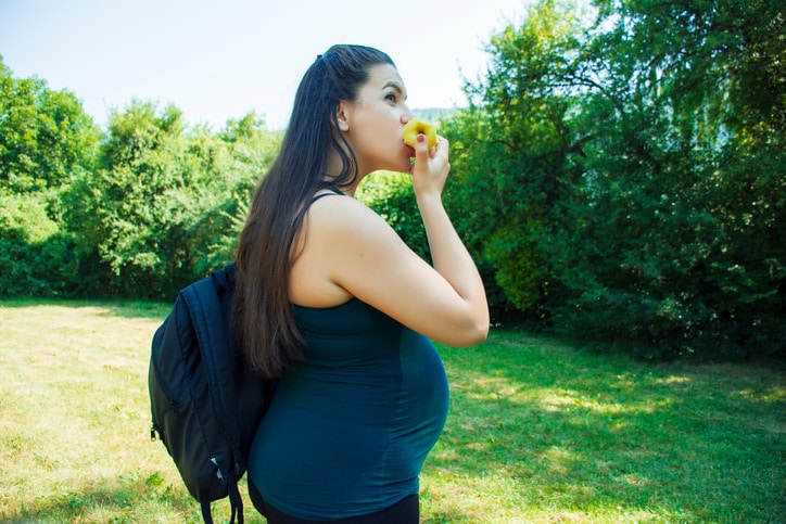 smiling pregnant woman eating an apple outdoors