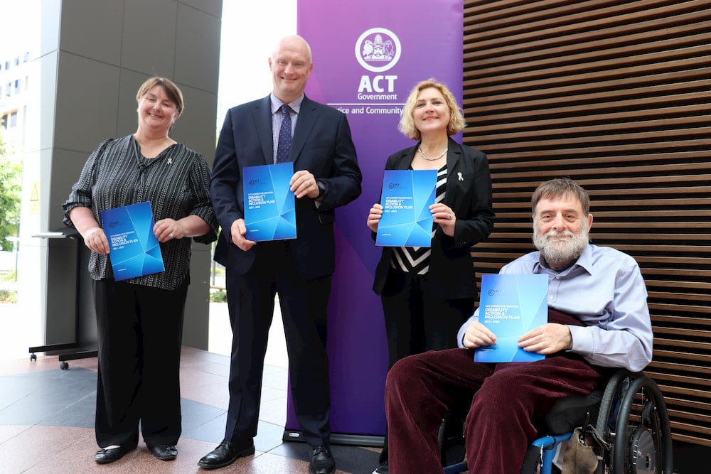Two women, one man standing and one man in wheelchair holding Disability Action and Inclusion Plan documents