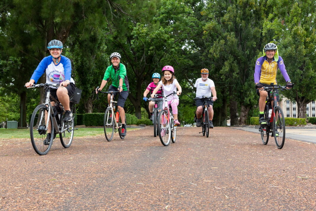 Six people in bikes, of all ages and abilities, ride toward the camera with smiles in their faces - they will all participate in the Big Canberra Bike Ride