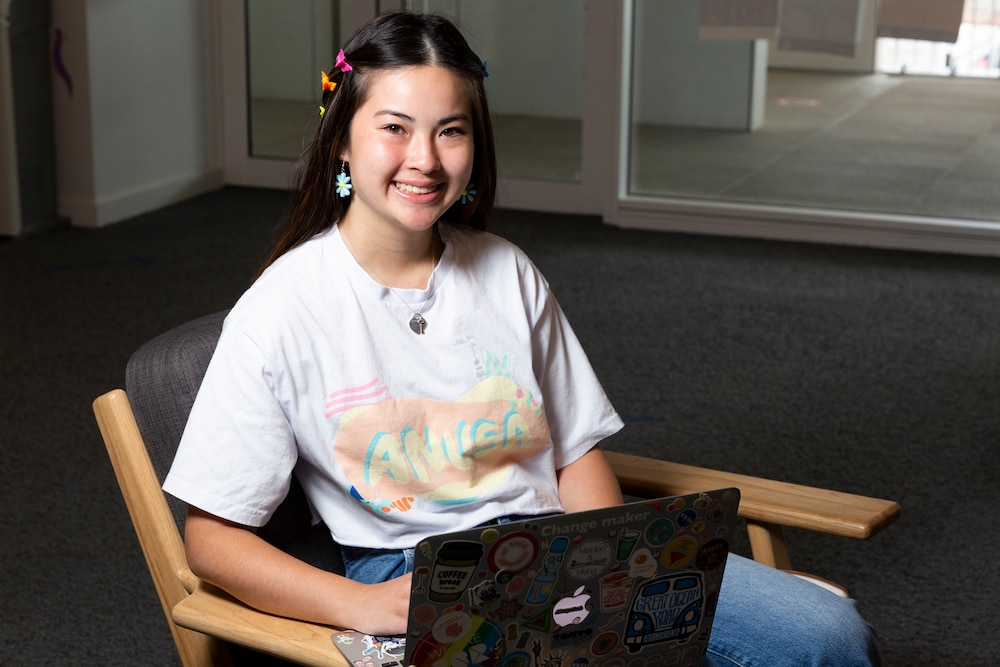 Madeleine Chia is pictured sitting in a wooden chair with her laptop open, wearing a white shirt and butterfly clips in her hair. She has experience in insecure work.
