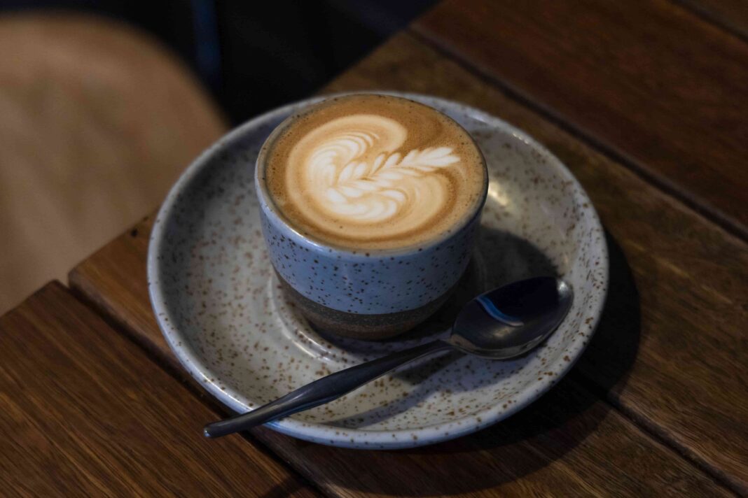 latte art on a coffee in a speckled stoneware cup on a stoneware saucer with teaspoon alongside