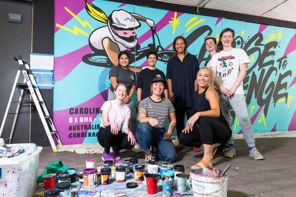 Caroline Buchanan crouches in front of a group of Erindale College students, and street artist Bohie kneels to her right. Art supplies litter the foreground. 