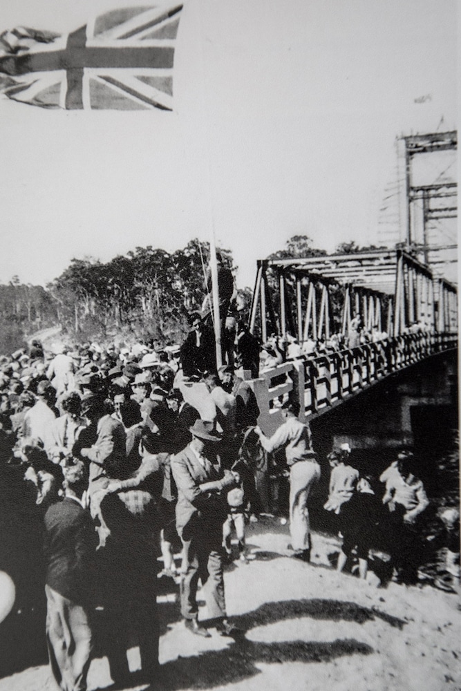 A black and white photo of a thick crowd of people in 1950s garb walking onto a bridge. 