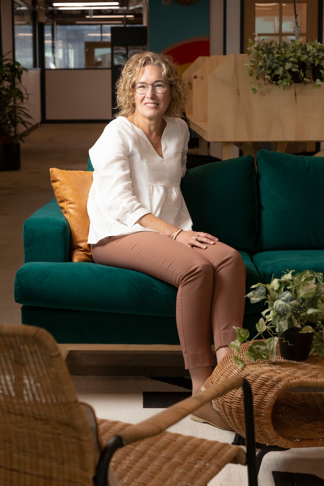 Louise wears glasses, a white top, light brown pants and sits on a green couch. She smiles toward the camera, and there is an ivy plant on the wicker table in front of her. 