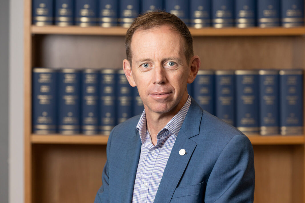 Unsmiling, ACT Attorney-General Shane Rattenbury wears a blue blazer and sits in front of a wooden bookshelf with blue books.