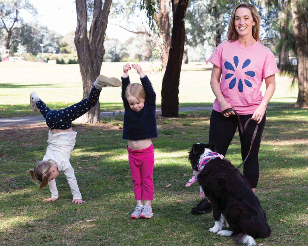 two little girls doing cartwheels as their mum in a pink t-shirt with dog on lead watches on, smiling