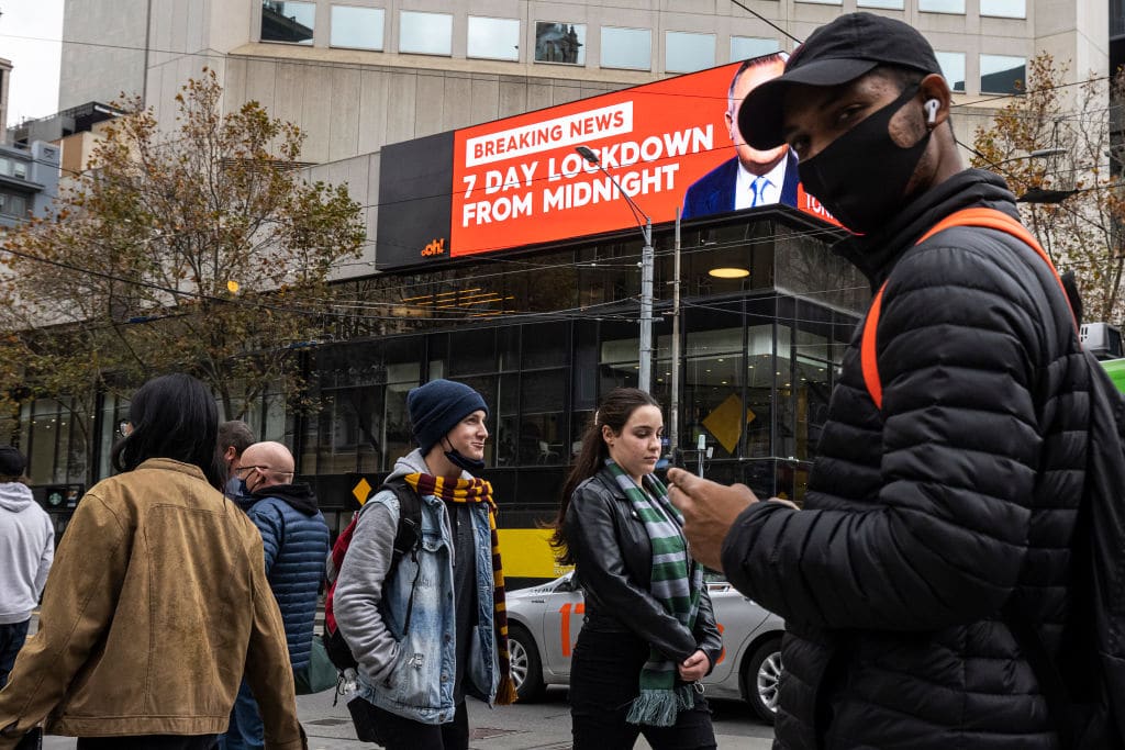 MELBOURNE, AUSTRALIA - MAY 27: People wearing face masks are seen crossing Bourke Street Mall in front of an LED sign in the background displaying news of the impending 7 day lockdown on May 27, 2021 in Melbourne, Australia. There are now 34 cases linked to a current cluster that spans several households and a workplace, with numerous exposure sites across many different parts of Melbourne and Bendigo. Victoria will go into a 7-day circuit breaker lockdown restrictions from 11:59 pm Thursday 27 May to 11:59 pm Thursday 3 June 2021. (Photo by Daniel Pockett/Getty Images)