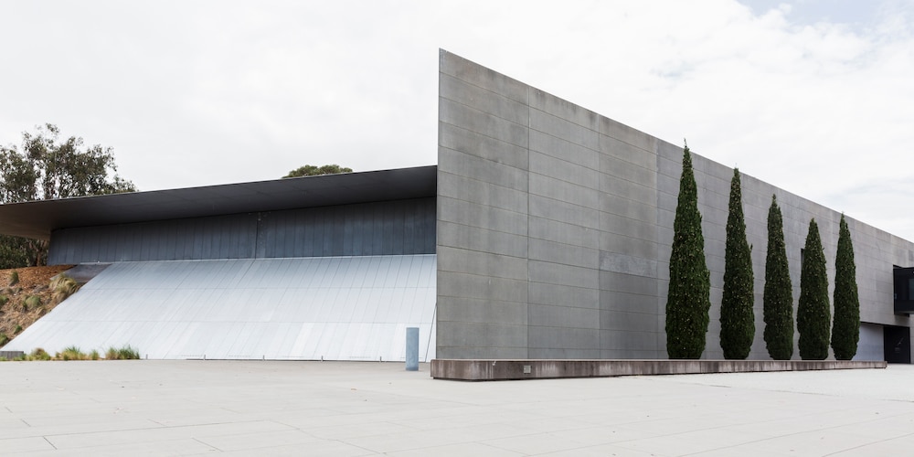Grey concrete exterior of Anzac Hall, an award-winning architectural space at the Australian War Memorial in Canberra, flanked by a row of conifers