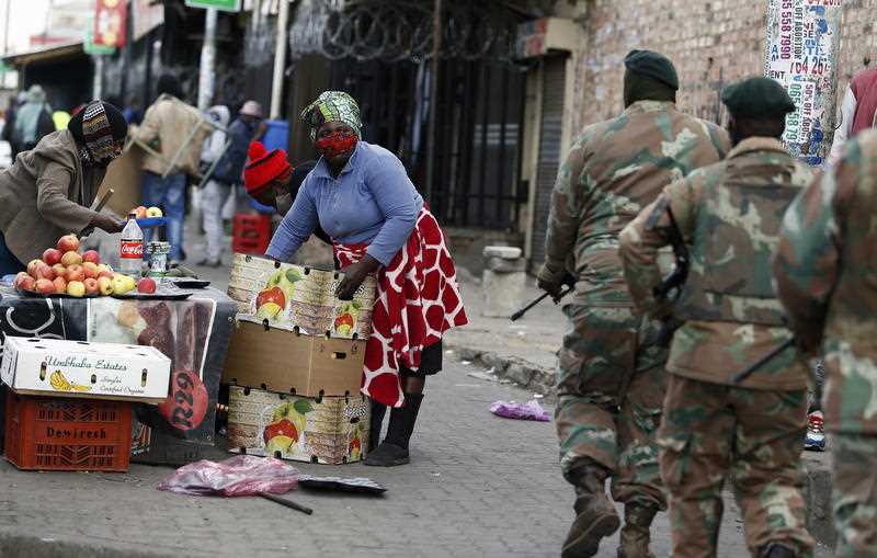 masked woman stocking fruit stall as troops patrol nearby in Alexandra Township, north of Johannesburg
