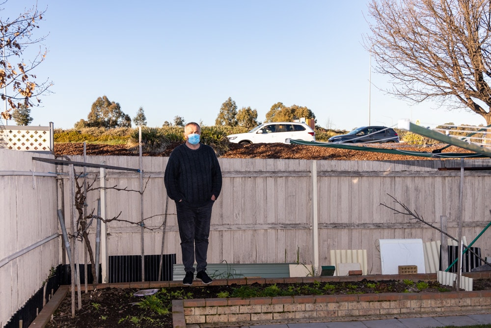 Man wearing mask standing in his backyard as traffic passes along a busy road beyond his fence