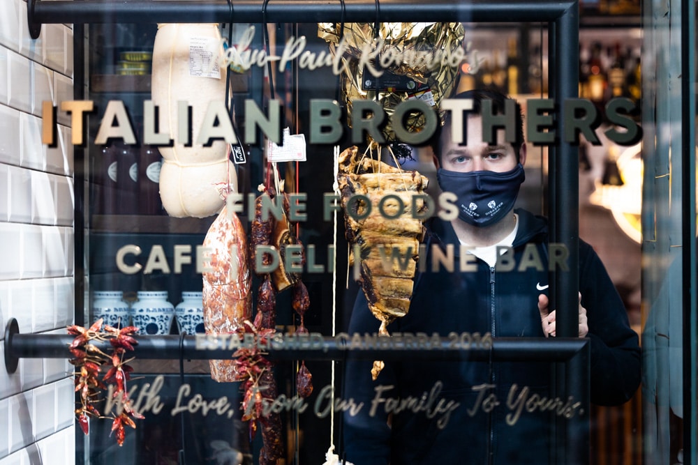 A Canberra business owner wearing face mask is seen through the window of his Italian deli in Manuka