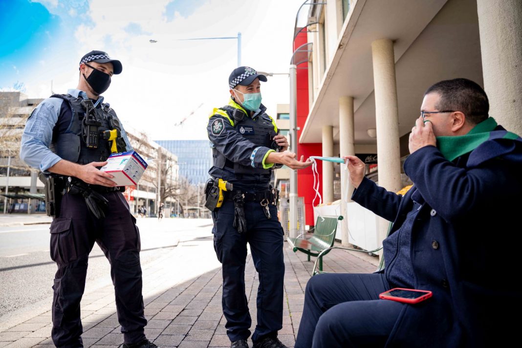 two ACT police officers are seen handing out a disposable face mask to a man sitting on a bench