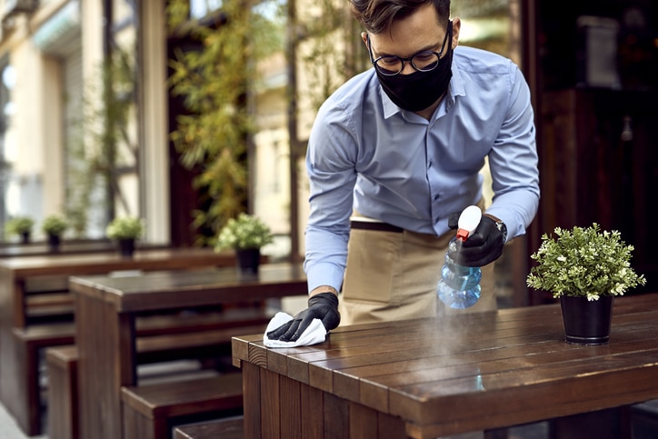 Waiter wearing protective face mask while disinfecting tables at outdoor cafe.