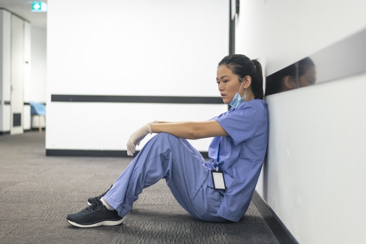 A female nurse of asian descent sits on the hospital hallway floor. The nurse appears exhausted and stressed after attending to patients with COVID-19 all day and night. She is emotionally drained and burnout from the long hours she has been working.