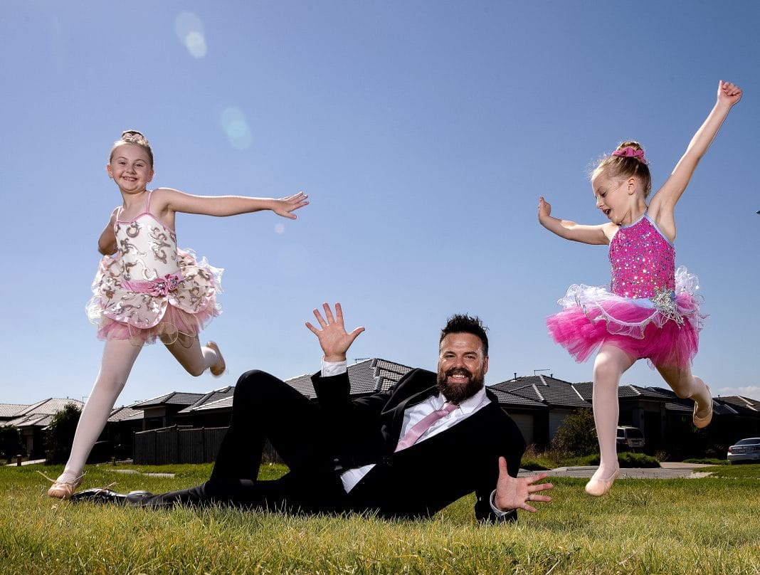 dressed in a suit with a pink tie, a dad poses with his two young ballerina daughters, aged 5 and 7