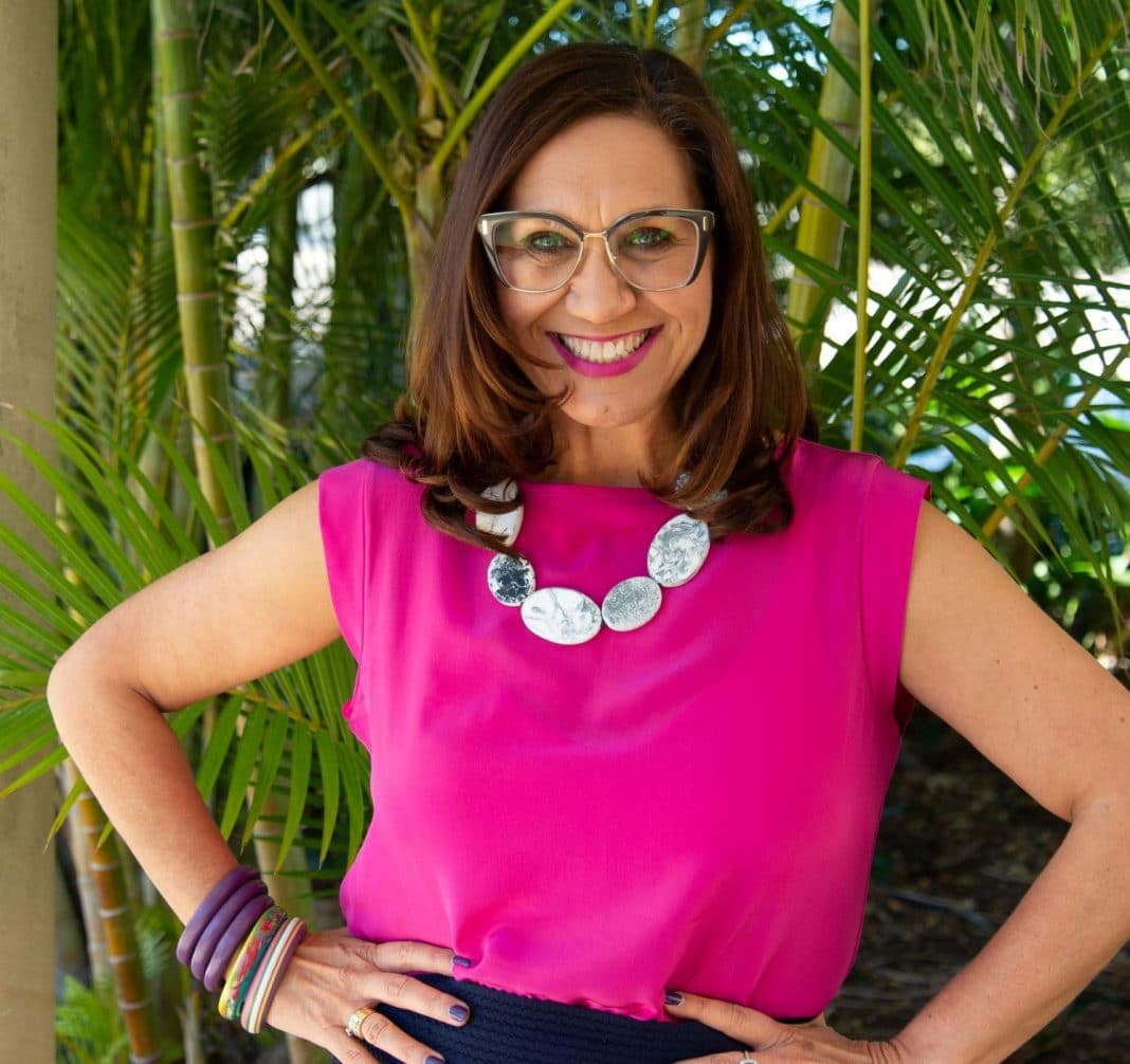 Indigenous author and academic Anita Heiss wearing, glasses, a bright pink top and chunky jewellery is seen smiling in a tropical garden