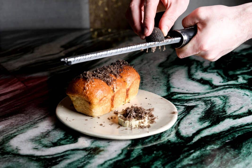 a hand is seen shaving black truffle over a mini loaf and round of butter on a white plate