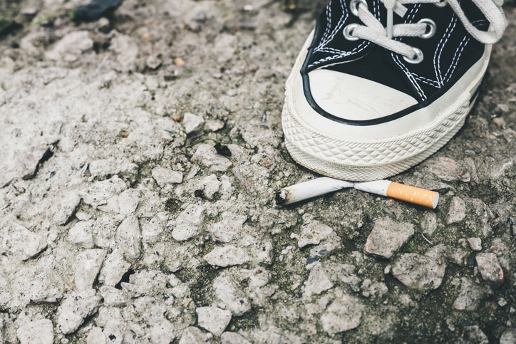 Closeup of a person’s foot wearing black sneakers shoes crushing a cigarette butt on asphalt. Quitting smoking concept
