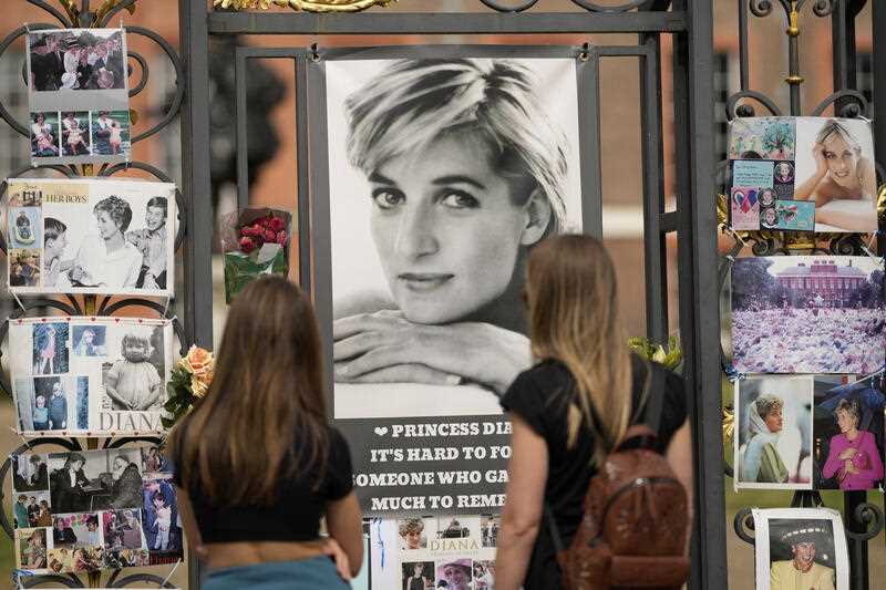 Two women look at portraits of Princess Diana and other remembrances displayed on the gates of Kensington Palace, in London, Tuesday, Aug. 30, 2022.