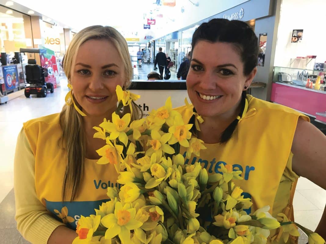 2 smiling female Cancer Council volunteers in a shopping centre selling Daffodils for Daffodil Day