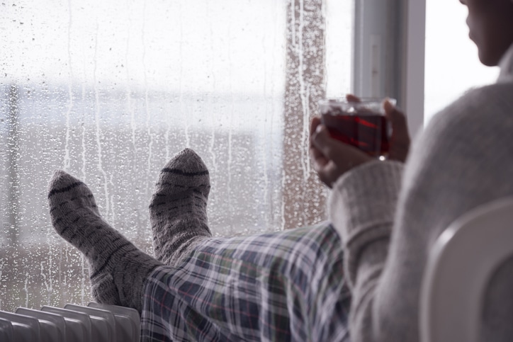 person with feet on heater drinking hot tea with condensation seen on window in background
