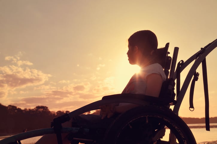 Teenage girl with cerebral palsy sits in a wheel chair watching the sunset by a river.