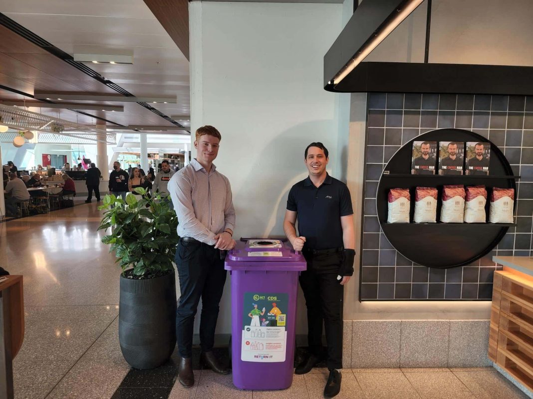 Two young men standing either side of a purple container deposit scheme recycling bin at Canberra Airport