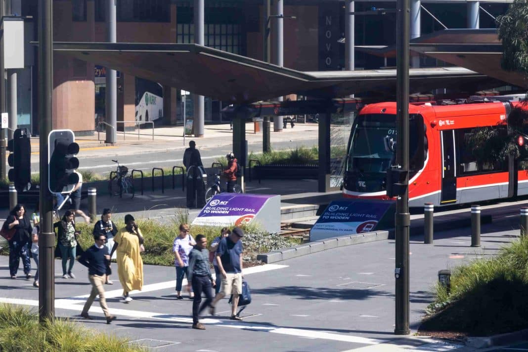 A red light rail vehicle is seen stopped at the terminus in Canberra city as pedestrians cross Northbourne Avenue
