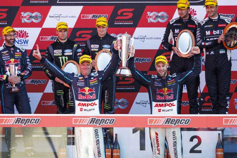 2 winning race car drivers on podium holding up Bathurst 1000 trophy with 5 other drivers standing behind them