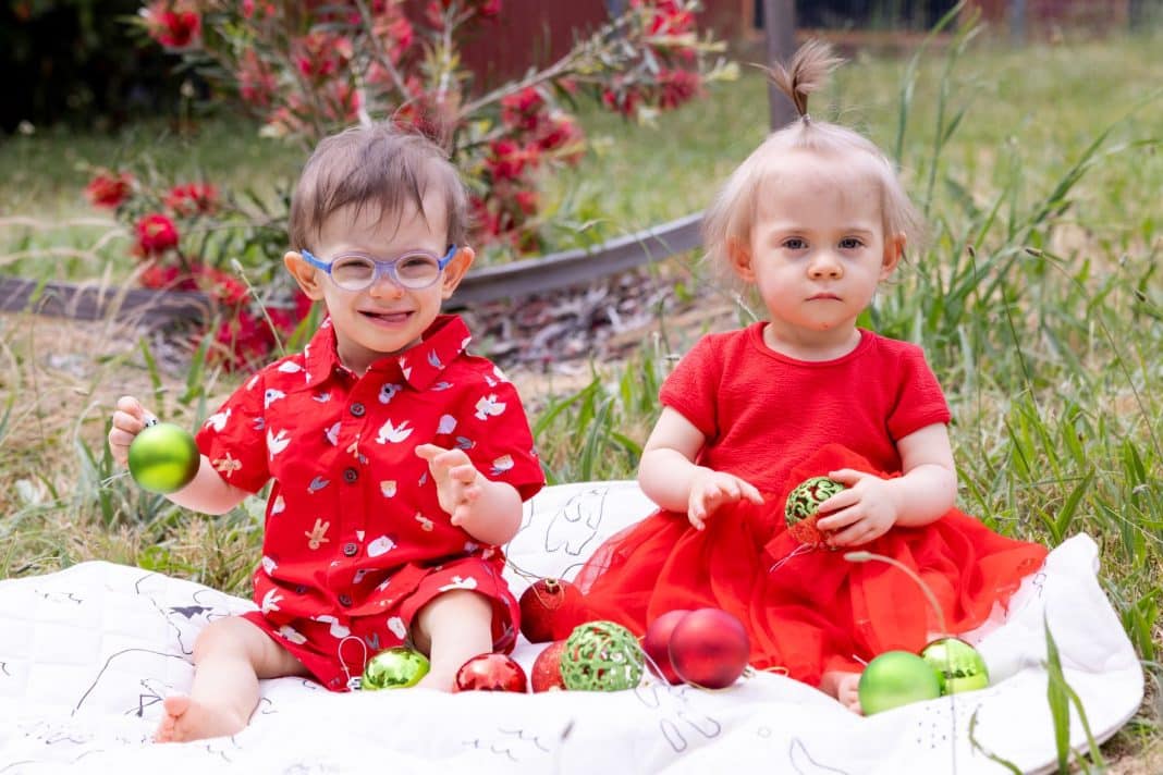 Cute boy and girl toddler twins in red outfits sitting on a picnic rug with red and green Christmas baubles