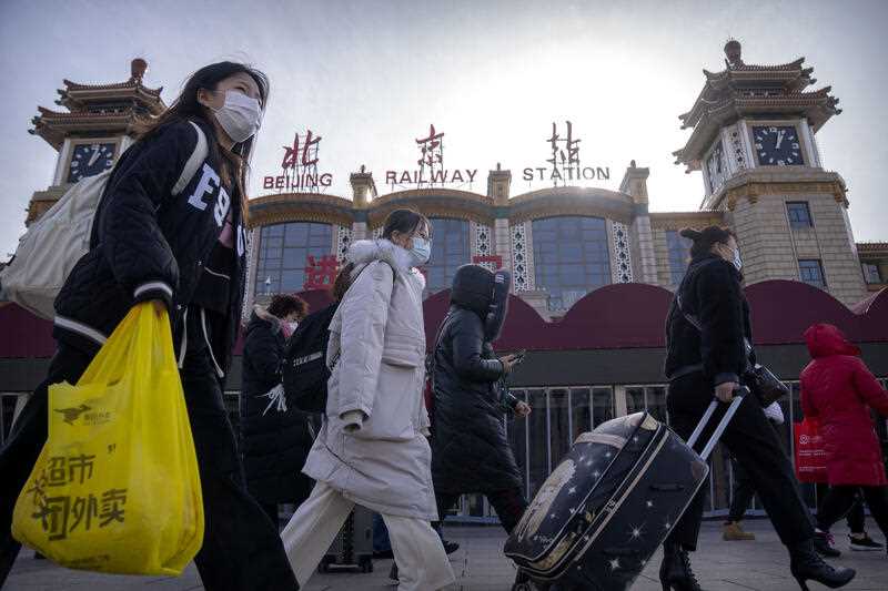 Travelers wearing face masks walk past the entrance of the Beijing Railway Station in Beijing, Saturday, Jan. 14, 2023