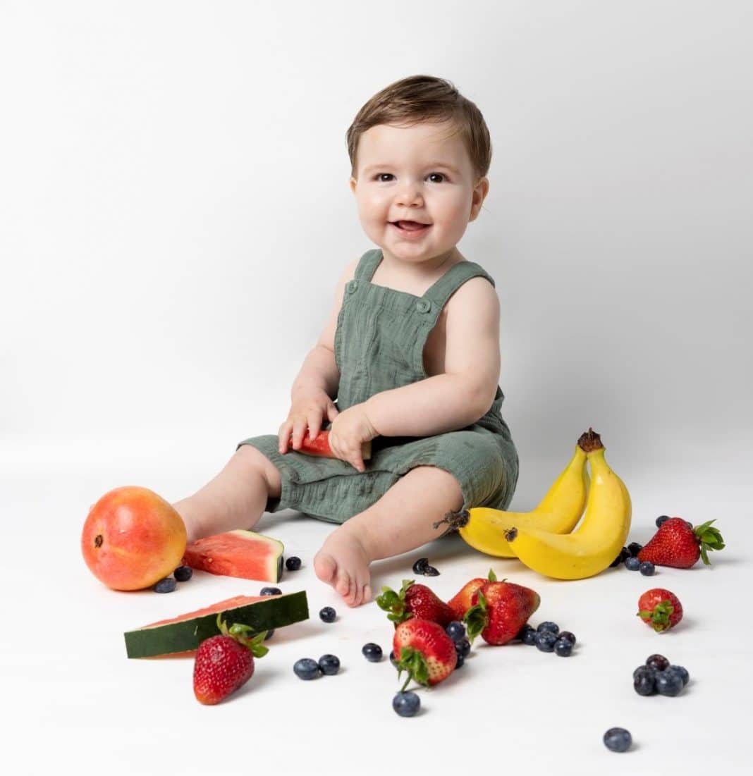 smiling one-year-old boy sitting on floor surrounded by fresh fruit
