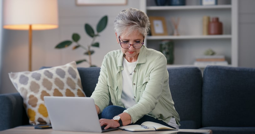 Senior woman typing on home laptop while reading notes
