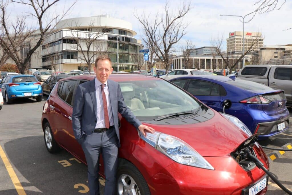 middle aged male politician in suit standing near an electric car in an outdoor carpark