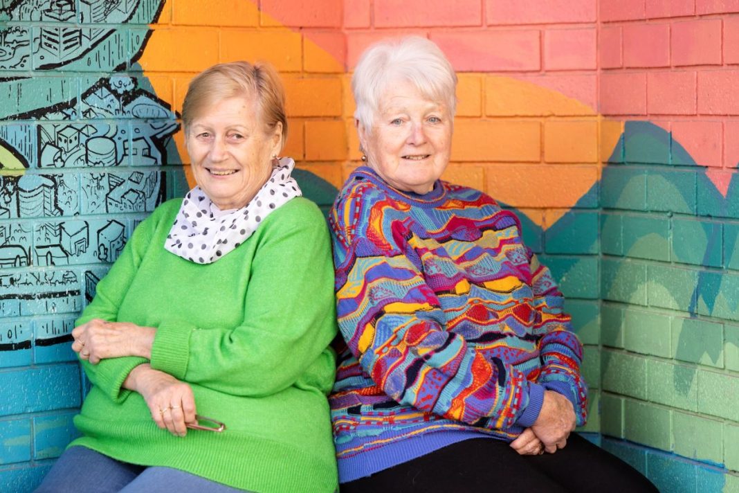 Two smiling senior women in colourful jumpers sitting on a bench in front of a colourful mural