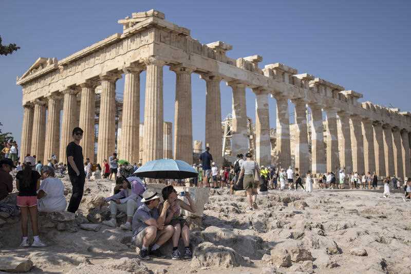 A tourist drinks water as she and a man sit under an umbrella in front of the five century BC Parthenon temple at the Acropolis hill during a heat wave, on July 13, 2023