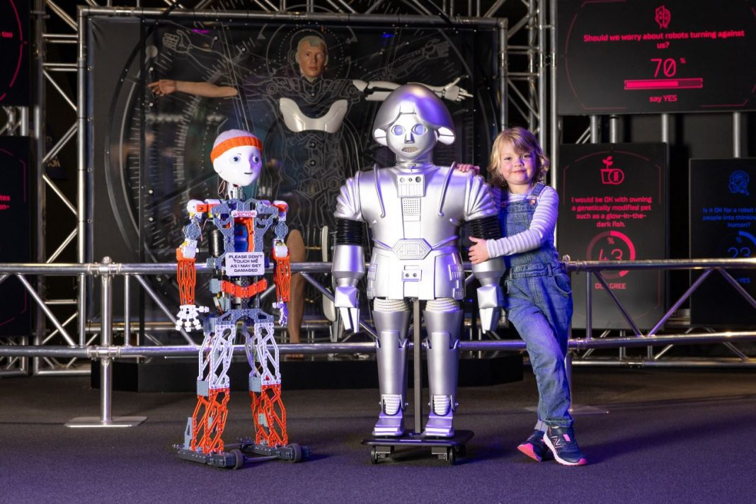 Cute 6 year old girl stands beside 2 different robots about the same size as her