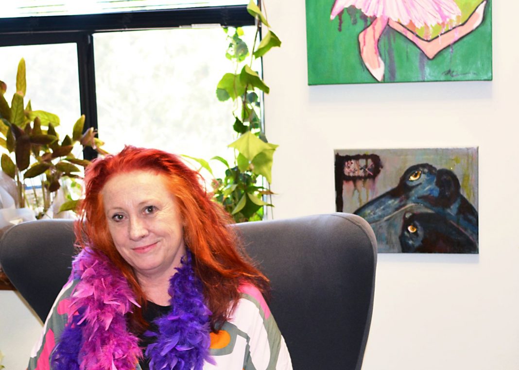 flamboyant female artist with red hair and purple and pink feather boa seated in front of colourful artworks