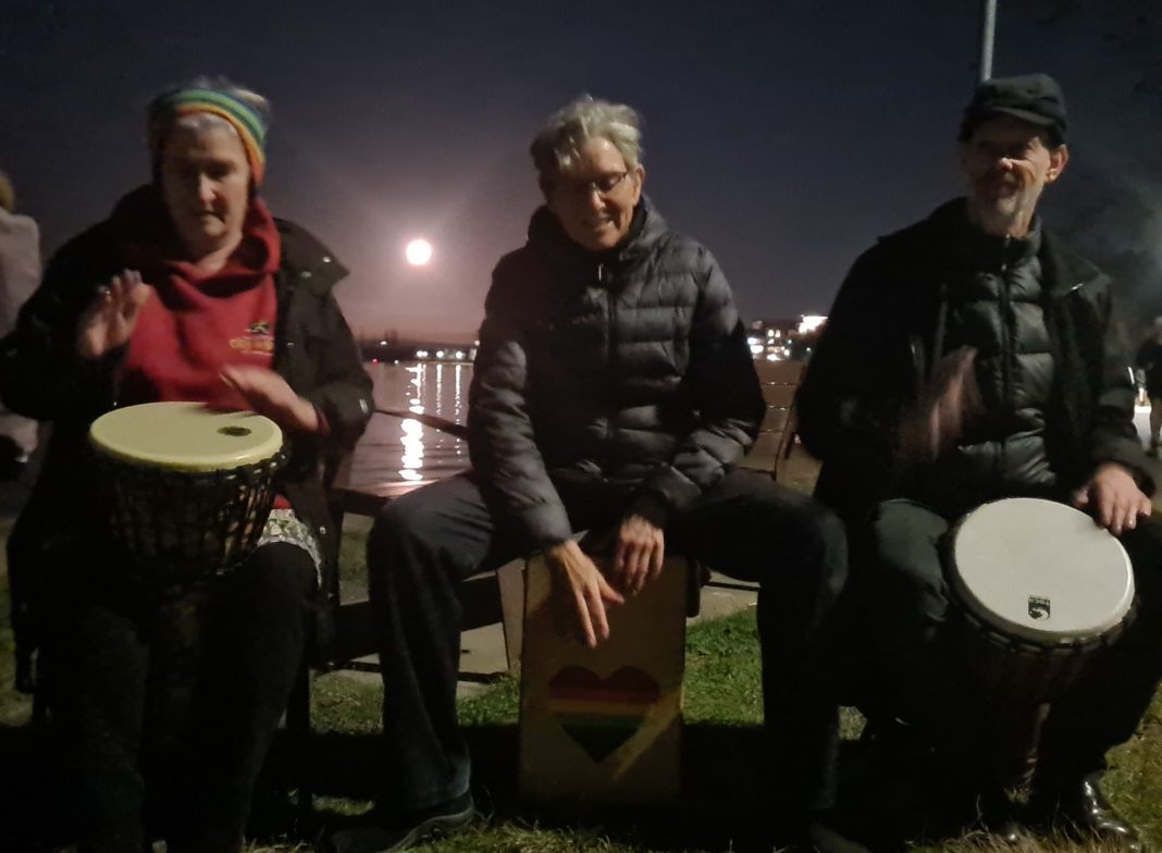 3 people drumming as full moon rises over Lake Burley Griffin