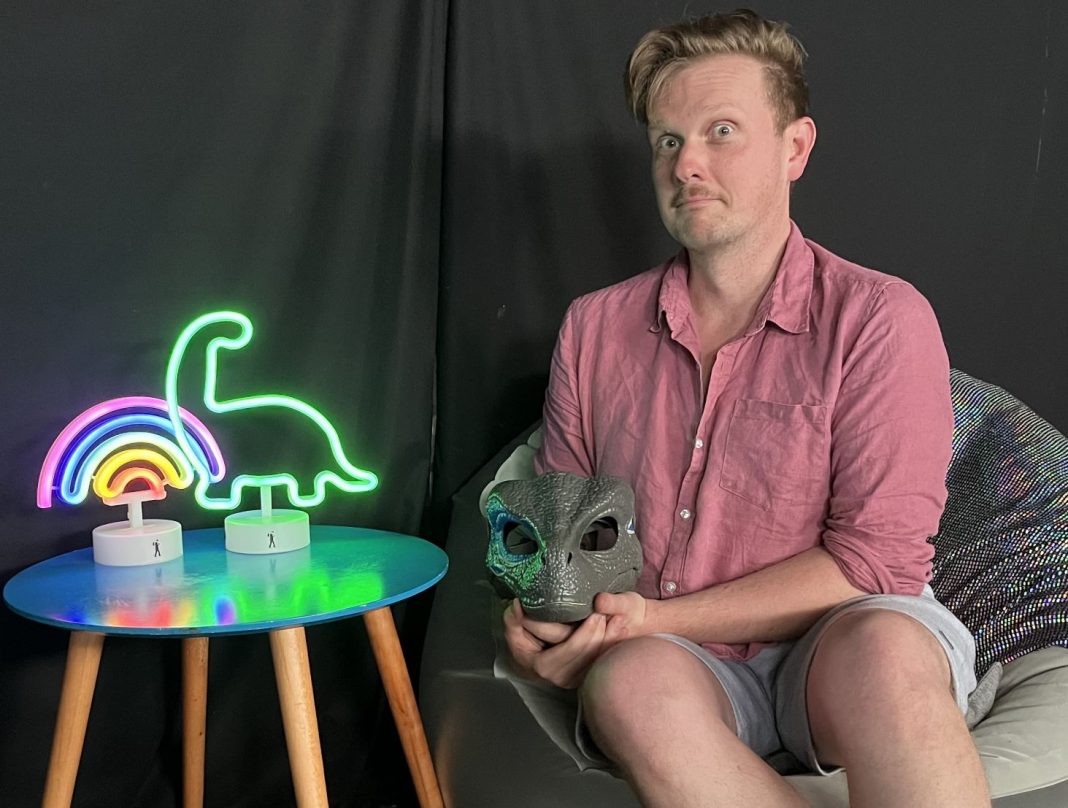 actor holding dinosaur mask sitting beside table with neon dinosaur and rainbow lights