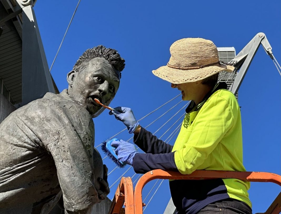 woman in hi-viz cleaning teeth of bronze Laurie Daley statue with a toothbrush
