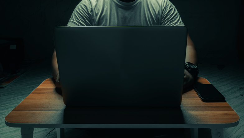An unidentifiable man in a t-shirt sits at a laptop in a dark room. with light shining down