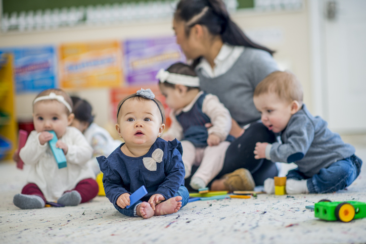 A group of babies are indoors in a daycare center. They are sitting on the floor and playing with their babysitter.